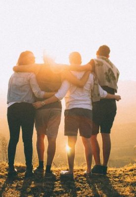 a group of people stand facing the sunset with arms around each other
