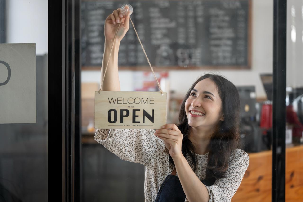 A smiling woman hangs an open sign up on a door