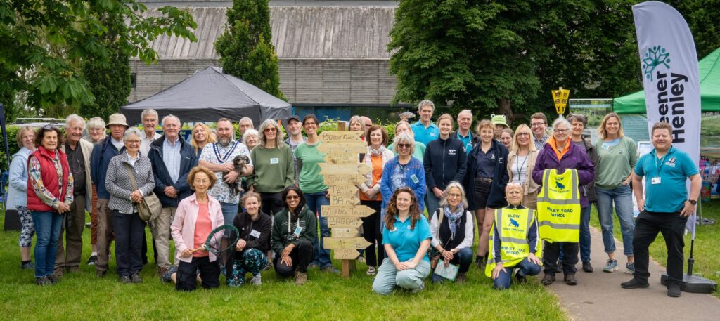 A large group of smiling happy people gather next to a Greener Henley banner, looking to camera