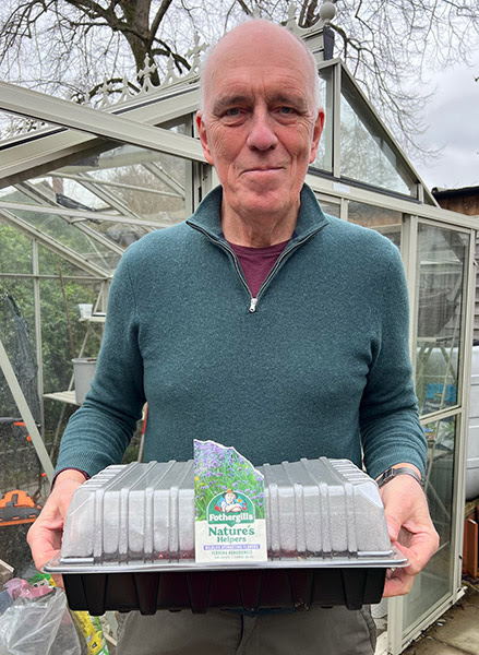 A man holds a tray of seedlings with a greenhouse in the background